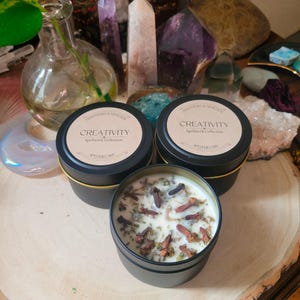 May include: Two black candle tins with gold lids. The candles are labeled "Creativity Spellwork Collection" and have a white wax with brown cloves inside.
