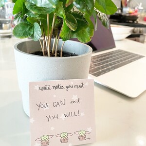 May include: A pink motivational note card with the text "write notes you must YOU CAN and YOU WILL!" and three cartoon characters. The card is sitting on a table next to a potted plant and a laptop.