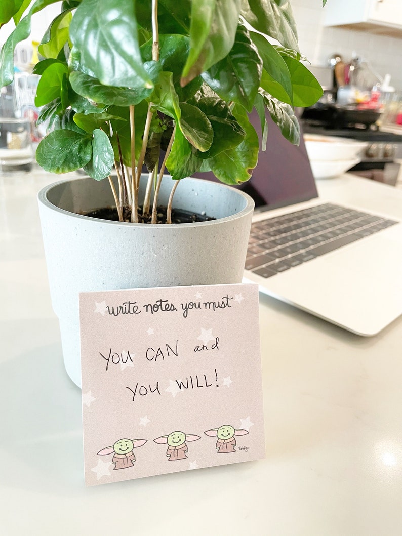 May include: A pink motivational note card with the text "write notes you must YOU CAN and YOU WILL!" and three cartoon characters. The card is sitting on a table next to a potted plant and a laptop.