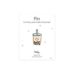 Puede incluir: Un pin de esmalte brillante con un diseño de té de burbujas sonriente. El pin está sobre una tarjeta blanca con la palabra "Pin" y la frase "I'm shiny and made of enamel!". La tarjeta también incluye la marca "tomkcy" y la dirección del sitio web.