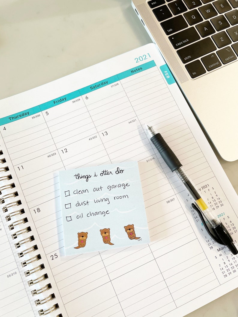 May include: A weekly planner with a blue and white colour scheme. The planner has a section for notes and a calendar for February 2021. A sticky note with the text "things i otter do" and a list of chores is attached to the planner. The list includes "clean out garage", "dust living room", and "oil change".