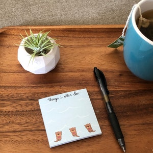 May include: A wooden tray with a white ceramic planter containing a green plant, a blue mug of tea, a black pen, and a blue sticky note with the text "Things I otter do" and three cartoon otters.