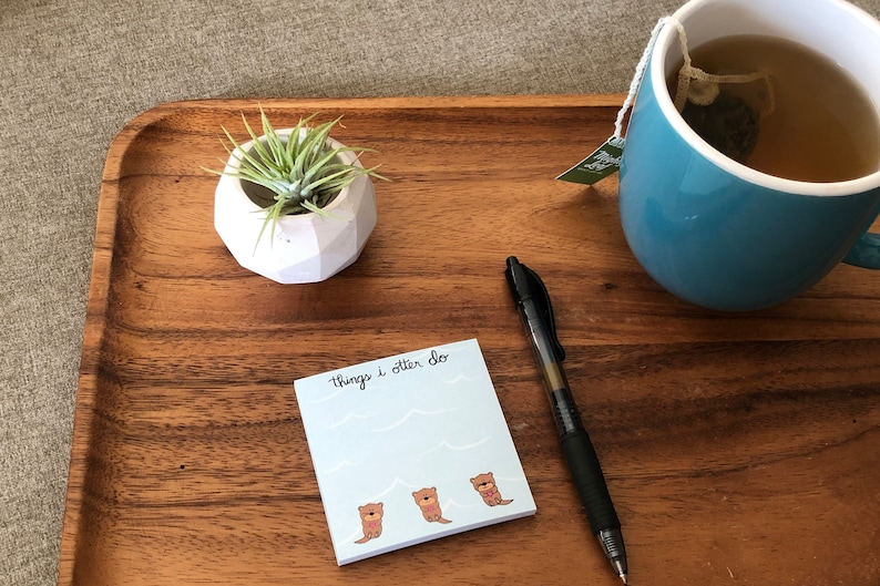 May include: A wooden tray with a white ceramic planter containing a green plant, a blue mug of tea, a black pen, and a blue sticky note with the text "Things I otter do" and three cartoon otters.