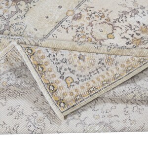 May include: A folded, vintage-style rug with a cream and beige color palette. The rug features a distressed, floral pattern with accents of brown and yellow. The edges of the rug are visible, showcasing a detailed border design.