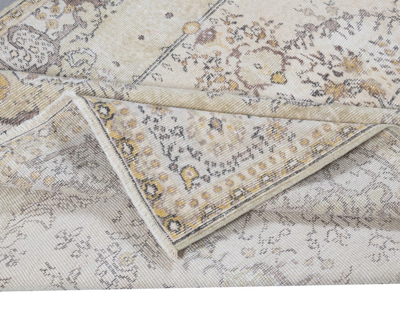 May include: A folded, vintage-style rug with a cream and beige color palette. The rug features a distressed, floral pattern with accents of brown and yellow. The edges of the rug are visible, showcasing a detailed border design.