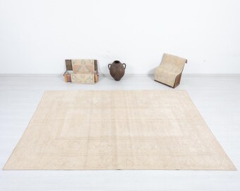 May include: A large, rectangular beige rug with a central geometric design. The rug is placed on a white floor, with a wooden chair, a decorative vase, and a wooden box with textiles in the background. The rug's dimensions are approximately 244 x 305 cm.