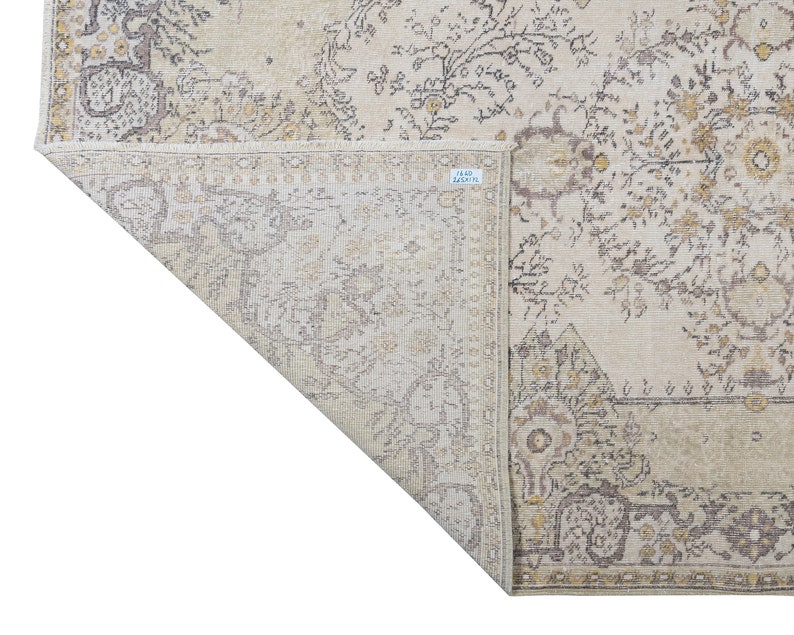 May include: Beige and gray area rug with an intricate floral pattern. The rug features a distressed, vintage look with a detailed border and corner designs. The rug is folded over, revealing the underside.