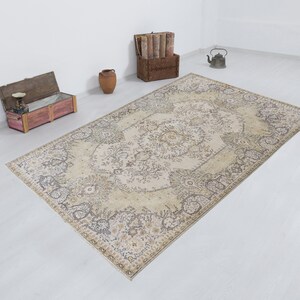 May include: A large, rectangular area rug with an intricate floral pattern in beige, cream, and muted brown tones. The rug's design features a central medallion and a detailed border. The rug is placed on a white floor, with decorative items in the background.
