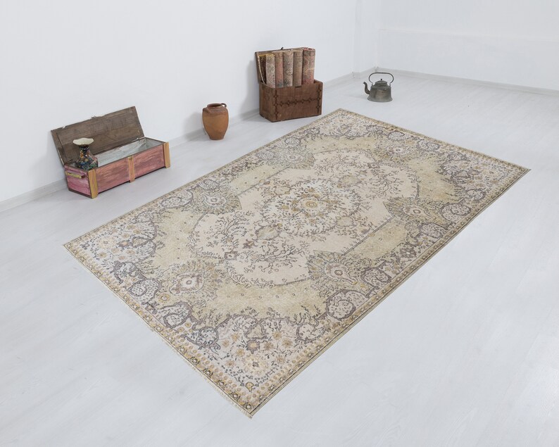 May include: A large, rectangular area rug with an intricate floral pattern in beige, cream, and muted brown tones. The rug's design features a central medallion and a detailed border. The rug is placed on a white floor, with decorative items in the background.
