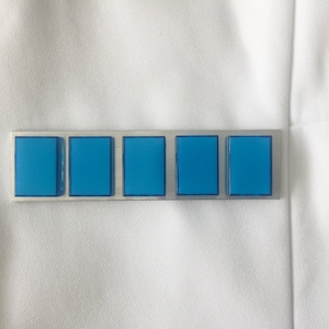 Puede incluir: Un alfiler de metal plateado con cinco incrustaciones rectangulares de color azul brillante. El alfiler está colocado sobre un fondo de tela blanca, lo que sugiere que podría estar sujeto a un uniforme o prenda. El diseño es simple y geométrico.