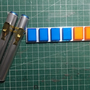 Peut inclure: Deux tubes argentés avec des fermoirs dorés et une rangée de cinq boutons carrés bleus et deux orange.