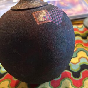 May include: A dark brown ceramic vase with a geometric design in pink, blue, and gold. The vase has a textured surface and is sitting on a colorful fabric.