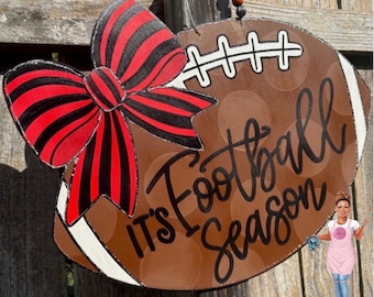 May include: A brown wooden football-shaped door hanger featuring a large red and black striped bow, white laces, and the black script text "It's Football Season". A small illustration of a person in a pink apron is attached to the lower right.