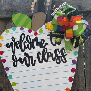 May include: Wooden apple-shaped sign for a classroom, featuring the words "welcome to our class" in black script. The sign has a colourful polka dot border and a decorative bow with green, black, and rainbow ribbons. A green and black striped leaf is attached.