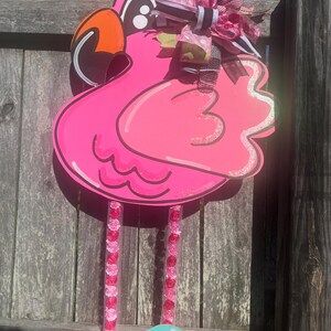 May include: Pink flamingo door hanger with a black and white bow, pink and white polka dot bow, and green and white bow. The flamingo has a black beak and orange inside the beak. The flamingo has pink and white beaded legs and is wearing orange and turquoise flip flops.
