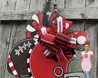 May include: A decorative door hanger shaped like a red football helmet and a brown football, accented with a large multi-layered bow featuring red, black, and white ribbons with paw prints and the text GEORGIA printed on the fabric strips.