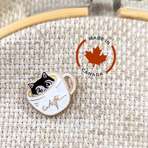 May include: A gold-coloured enamel pin featuring a black cat peeking out of a white coffee mug with the word "Cafe" written on it. The pin is on a beige fabric background. Made in Canada.
