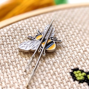 May include: A silver bee-shaped needle minder with yellow, black, and white enamel detail, holding two silver needles. The bee is resting on a beige cross-stitch fabric, with a wooden embroidery hoop in the background. The needles are for cross-stitch or embroidery.