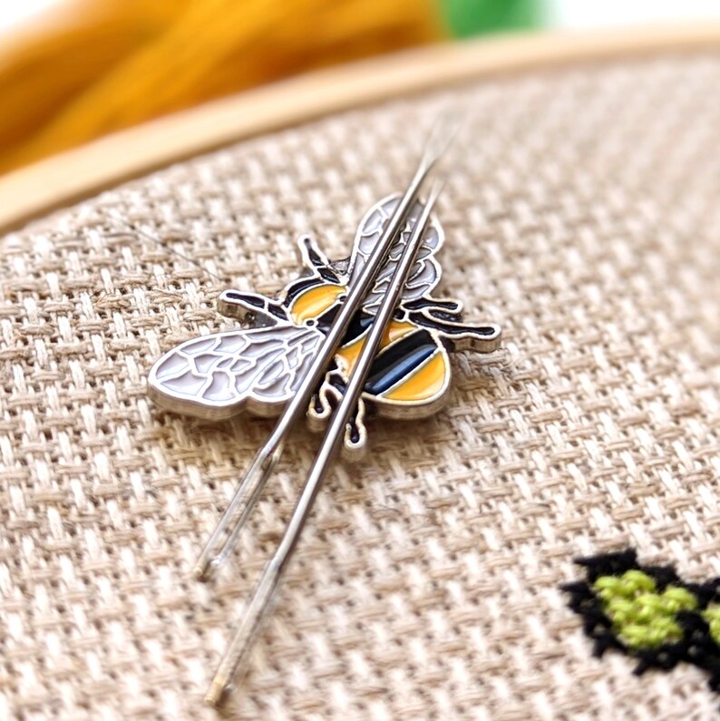 May include: A silver bee-shaped needle minder with yellow, black, and white enamel detail, holding two silver needles. The bee is resting on a beige cross-stitch fabric, with a wooden embroidery hoop in the background. The needles are for cross-stitch or embroidery.