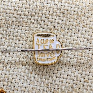 May include: A white enamel pin shaped like a mug with gold trim and lettering that reads A CUP OF TEA SOLVES EVERYTHING. A long, thin, silver-coloured metal sewing needle is positioned horizontally across the centre of the mug-shaped pin.