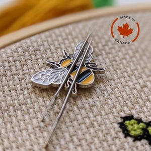 May include: A bee-shaped needle minder with two needles on a cross-stitch project. The bee has silver wings, a yellow and black body, and is marked "Made in Canada". The needles are silver and are positioned near the bee.