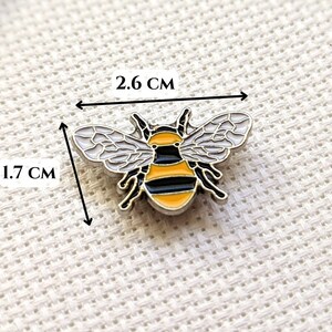 May include: A bee-shaped enamel pin, measuring 1.7 cm by 2.6 cm. The pin has a black and yellow striped body, white wings with black outlines, and black antennae. The pin is set against a textured white background.