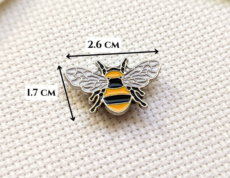 May include: A bee-shaped enamel pin, measuring 1.7 cm by 2.6 cm. The pin has a black and yellow striped body, white wings with black outlines, and black antennae. The pin is set against a textured white background.