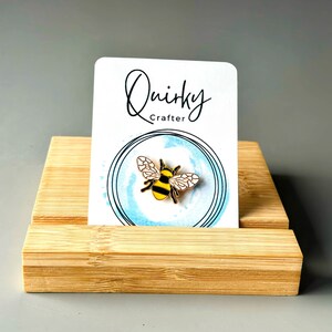 May include: A gold-coloured bee pin with black and yellow stripes, presented on a white card with the text "Quirky Crafter". The card is set against a light blue watercolour circle and rests on a bamboo display stand.