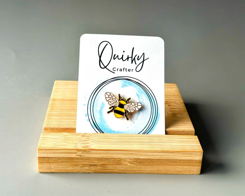 May include: A gold-coloured bee pin with black and yellow stripes, presented on a white card with the text "Quirky Crafter". The card is set against a light blue watercolour circle and rests on a bamboo display stand.