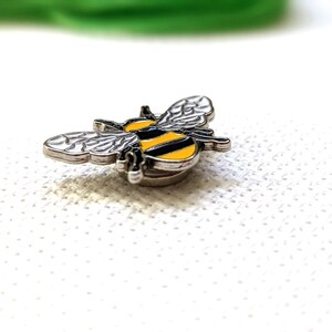 May include: A miniature bee-shaped brooch with a silver border. The bee has black and yellow stripes on its body and silver wings. The brooch is set against a white, textured surface.