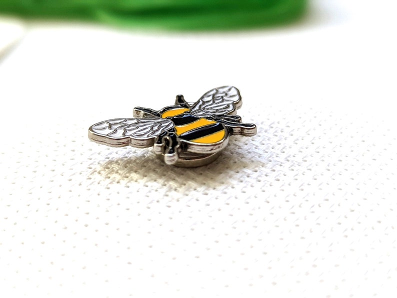 May include: A miniature bee-shaped brooch with a silver border. The bee has black and yellow stripes on its body and silver wings. The brooch is set against a white, textured surface.