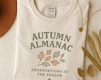 May include: Cream-colored t-shirt with the text "AUTUMN ALMANAC" in green and a leaf graphic. Below the text is "OBSERVATIONS OF THE SEASON". A gold necklace and dried leaves are also visible.