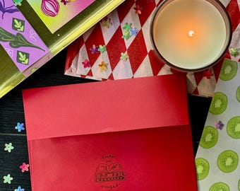 May include: A bright red envelope featuring a small black ink illustration of an open book with the text "into THE pages" centered on the flap. The envelope is surrounded by colorful confetti, a gold tray, a lit candle, and patterned paper.