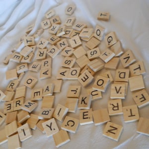 Pode incluir: Um conjunto de peças de letras de madeira, comumente usadas no jogo Scrabble, estão espalhadas sobre uma superfície de tecido branco. As peças estão todas em letras maiúsculas, e algumas das letras são repetidas.