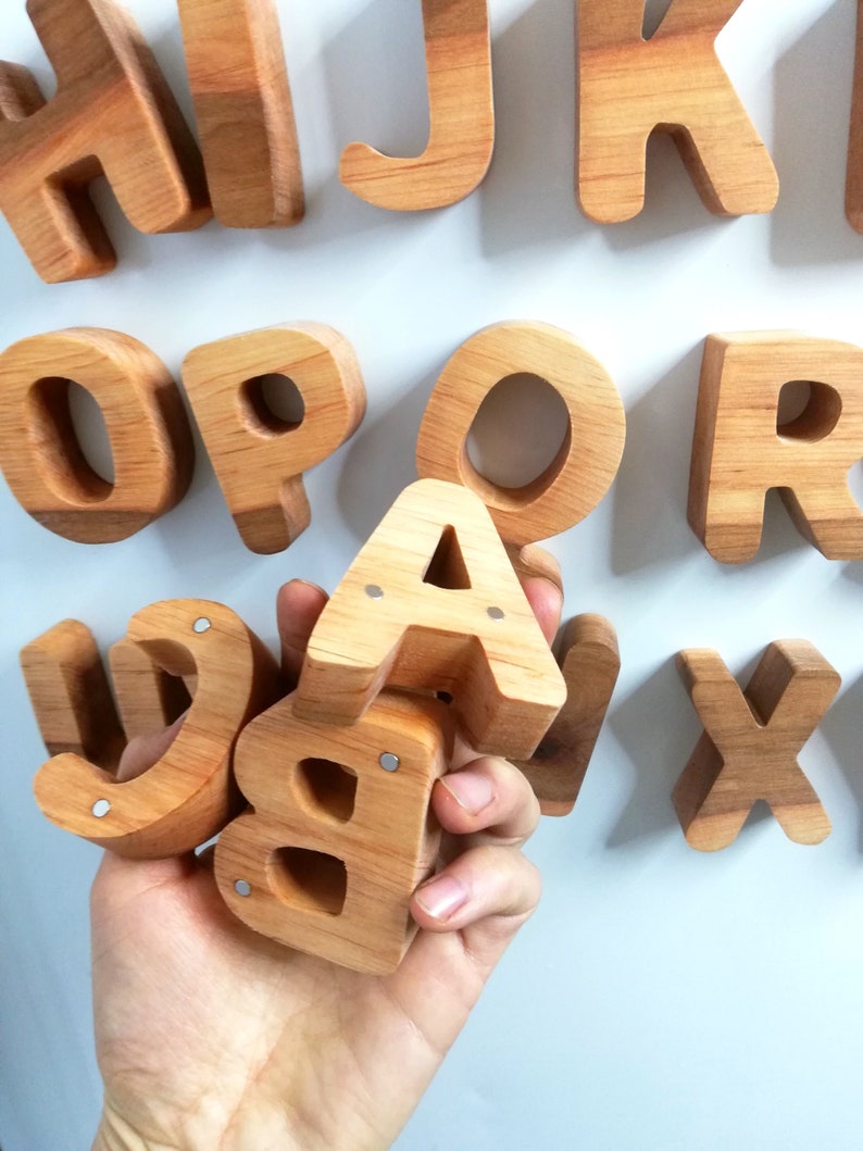 May include: Wooden alphabet blocks in various letters, including A, B, C, J, K, O, P, R, U, X, and Y. The blocks are made of light brown wood and have a natural finish. Some of the blocks have small metal dots on the back.