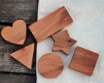 May include: Set of six wooden shapes in natural wood tones, including a heart, star, circle, square, and two triangles. The shapes are arranged on a white fabric surface.