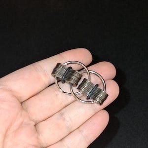 May include: Two silver keychains interlocked, each with a bundle of small, rectangular metal pieces held together by a black rubber band. The keychains are circular and are held in a hand against a dark background.