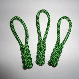 May include: Three green and black paracord zipper pulls are displayed against a white background. Each pull features a looped top and a braided bottom, designed for easy gripping and use on jackets, bags, or other items.
