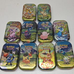 May include: A collection of ten Pokémon Trading Card Game tins. Each tin features a different Pokémon character illustration on a green and blue background. The tins are rectangular with rounded edges and a gold metallic finish.