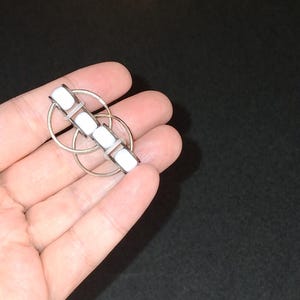 May include: A silver-toned brooch with a geometric design. The brooch features a series of four white rectangular stones set in a linear pattern, connected by a silver metal frame. Two interlocking circles are also part of the design.