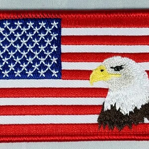 Könnte beinhalten: Gestickter Aufnäher mit der amerikanischen Flagge und einem Weißkopfseeadler auf der rechten Seite. Die Flagge ist rot, weiß und blau mit 13 Streifen und 50 Sternen. Der Adler ist weiß mit einem gelben Schnabel und braunen Federn.