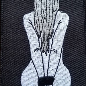 Op de afbeelding: Wit geborduurd beeld van een persoon met lang haar dat achter de rug is vastgebonden, zittend met gebonden handen voor zich.