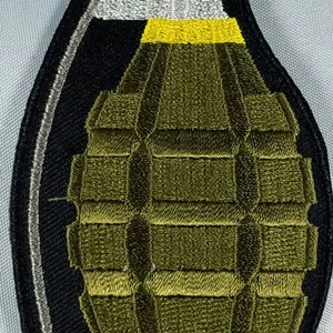 May include: A green and black embroidered patch of a grenade with a yellow pull pin.