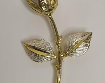 May include: A rose-shaped brooch featuring a polished gold-tone stem and thorns, with silver-tone petals and leaves. The metal surfaces exhibit a textured, etched finish on the leaves and petals, contrasting with the smooth, reflective gold-tone stem.