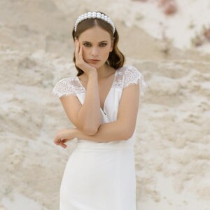 May include: A woman wearing a white wedding dress with a lace bodice and a slit in the skirt. She is standing in a sandy desert landscape and has a pearl headband in her hair.