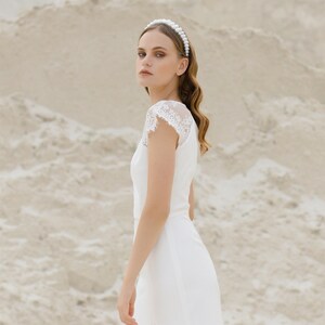 May include: A woman in a white wedding dress with a pearl headband stands in a sandy landscape. The dress has a lace bodice and a slit up the side.
