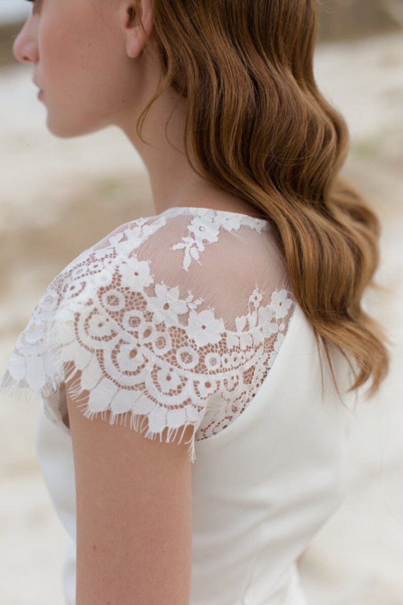 May include: A white wedding dress with a lace overlay on the shoulders. The dress has a simple, elegant design.