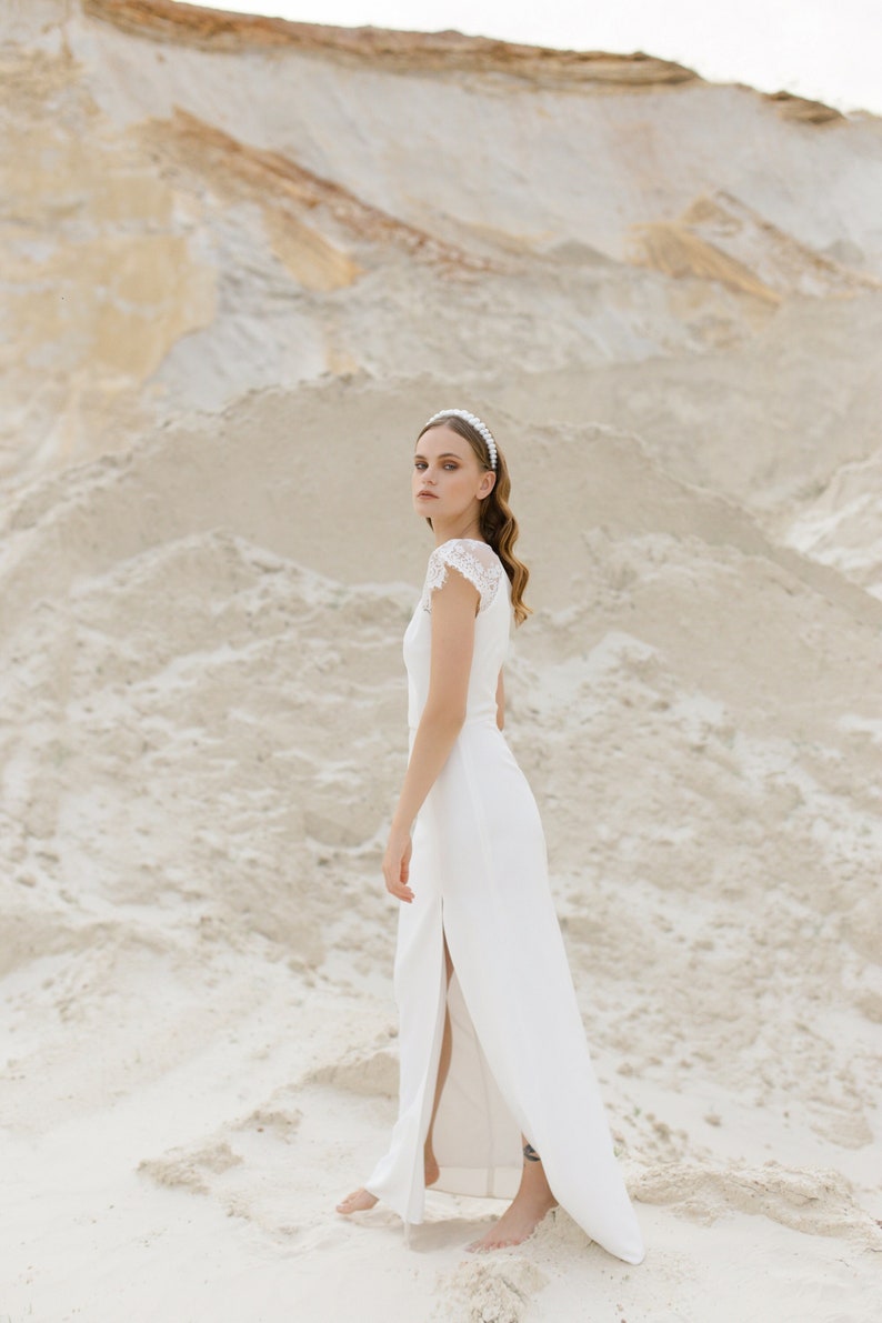 May include: A woman in a white wedding dress with a pearl headband stands in a sandy landscape. The dress has a lace bodice and a slit up the side.