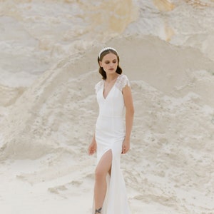 May include: A woman in a white wedding dress with a lace bodice and a slit up the leg stands in a sandy area. She is wearing a pearl headband and has her hair pulled back.