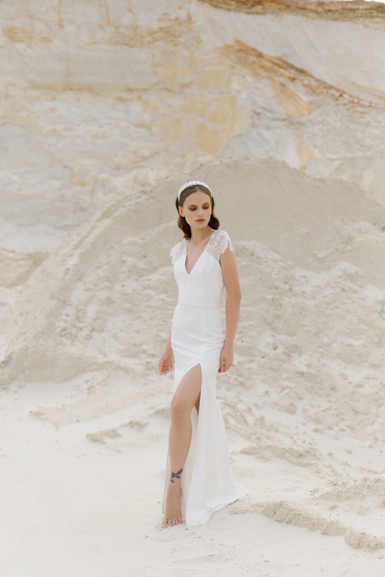 May include: A woman in a white wedding dress with a lace bodice and a slit up the leg stands in a sandy area. She is wearing a pearl headband and has her hair pulled back.